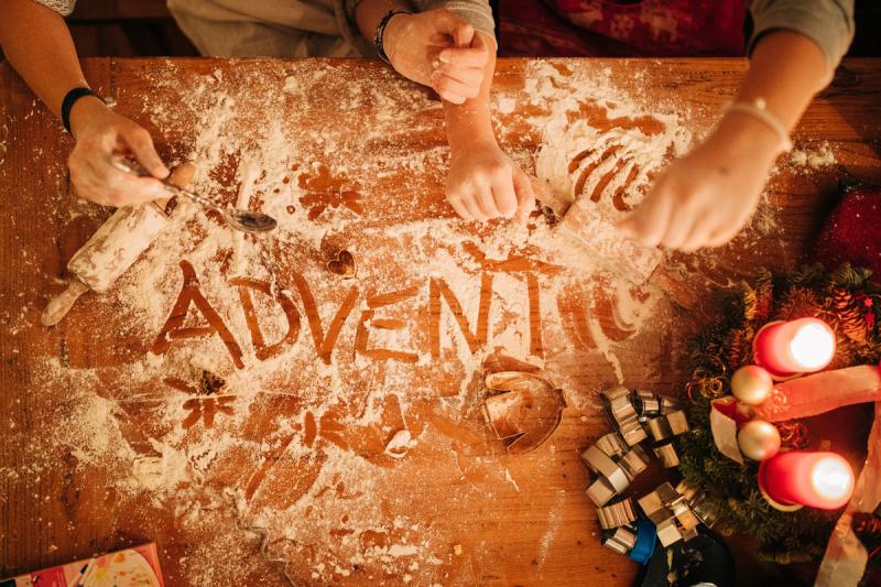 Man sieht Kinderhände, die beim Backen im Mehl auf einer braunen Tischplatte das Wort Advent geschrieben haben. 